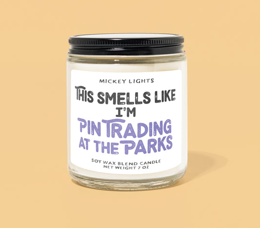 Close-up photo of a Disney-inspired candle with the label 'This Smells Like I’m Pin Trading at the Parks.' The candle features scent notes of sparkling citrus, cinnamon powder, macintosh apple, spicy rose, clove buds, and sugared vanilla. Background includes colorful Disney pin trading lanyards and pins, capturing the magic and nostalgia of Disney Parks.